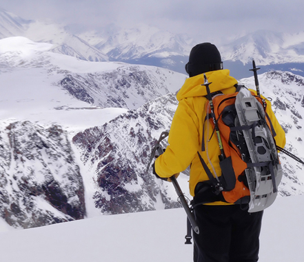 A hiker in a bright yellow jacket standing at a snowy vantage point overlooking a mountain range, with ski poles and a backpack, contemplating the vast, wintry landscape.