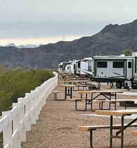 A row of RVs lines a gravel campground with picnic tables and a white fence, set against rugged desert mountains.