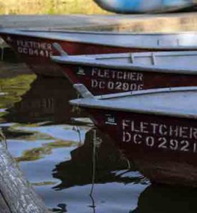 Three red rowboats labeled “FLETCHER” are moored side by side on calm water near a wooden dock in soft daylight.