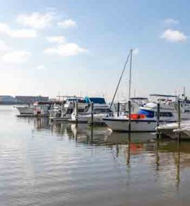 Motorboats and sailboats are docked in neat rows on calm water beneath a bright sky with scattered white clouds.