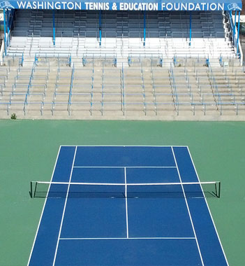 A bird's eye view of a tennis court with an empty spectator stand in the background, under the banner of the washington tennis & education foundation.