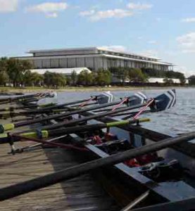 Rowing shells with paddles rest on a wooden dock by the water, with a large white-columned building in the distance.