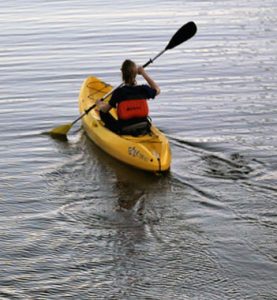 A person paddles a yellow kayak across rippling water, wearing a red life jacket and moving toward the open distance.