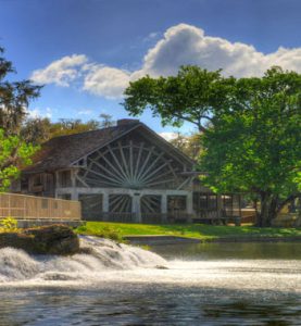 A serene lakeside retreat featuring a rustic building with distinctive architecture, surrounded by lush trees, under a blue sky scattered with clouds, with a cascading waterfall in the foreground.