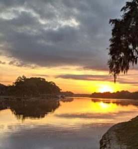 Tranquil sunset over a calm river, where the golden hues of the fading sun meet the gentle embrace of the water, framed by silhouettes of serene nature.