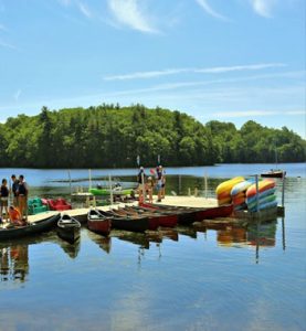 Colorful kayaks and canoes rest beside a dock on a calm lake, where people prepare to paddle under clear blue skies.