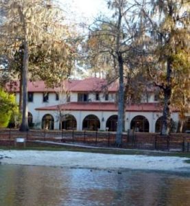 A tranquil view of a two-story building with a terracotta tiled roof, nestled among trees draped with spanish moss, overlooking a serene pond.