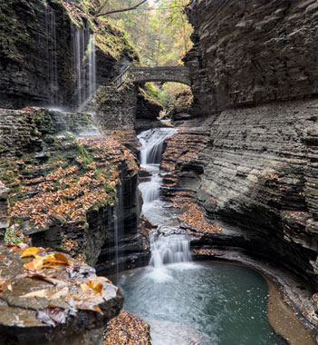 Waterfalls cascade down rocky cliffs in Watkins Glen, framed by autumn leaves and a stone arch bridge above.