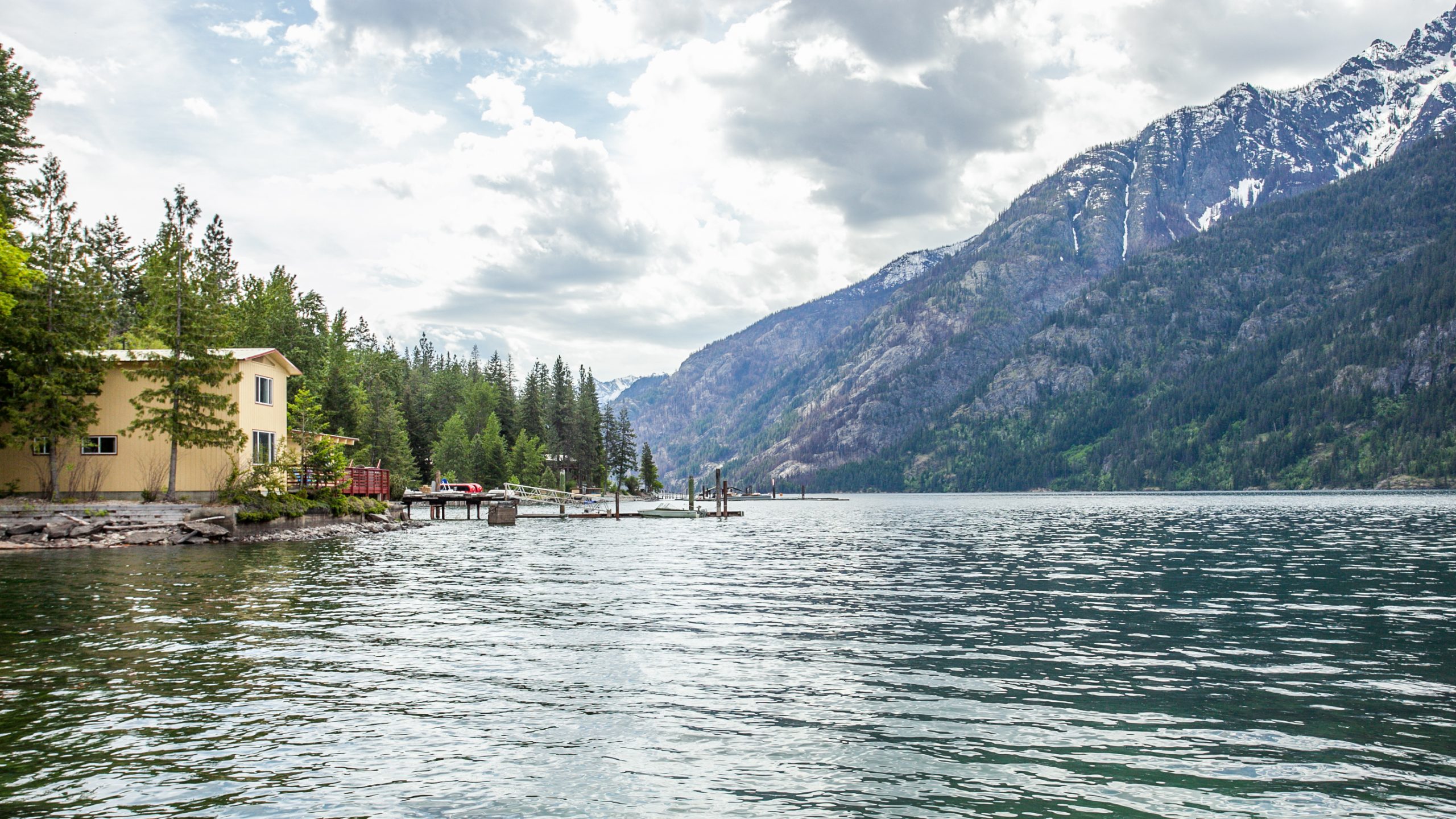 A tranquil lakeside house surrounded by dense evergreen forests and towering, snow-capped mountains.