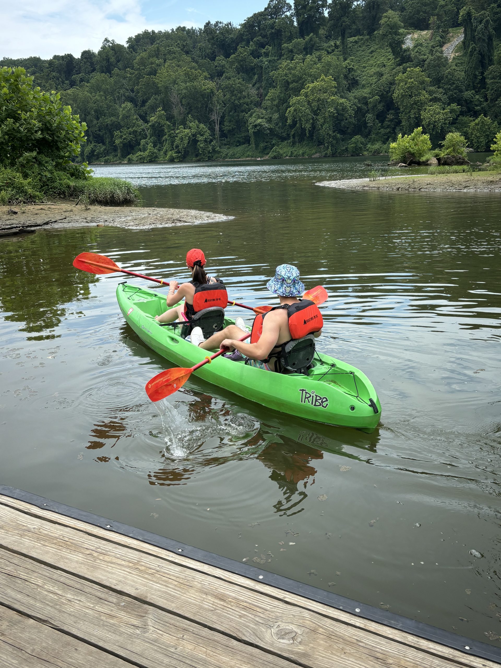 Two people in a green tandem kayak paddling away from a dock on calm water.