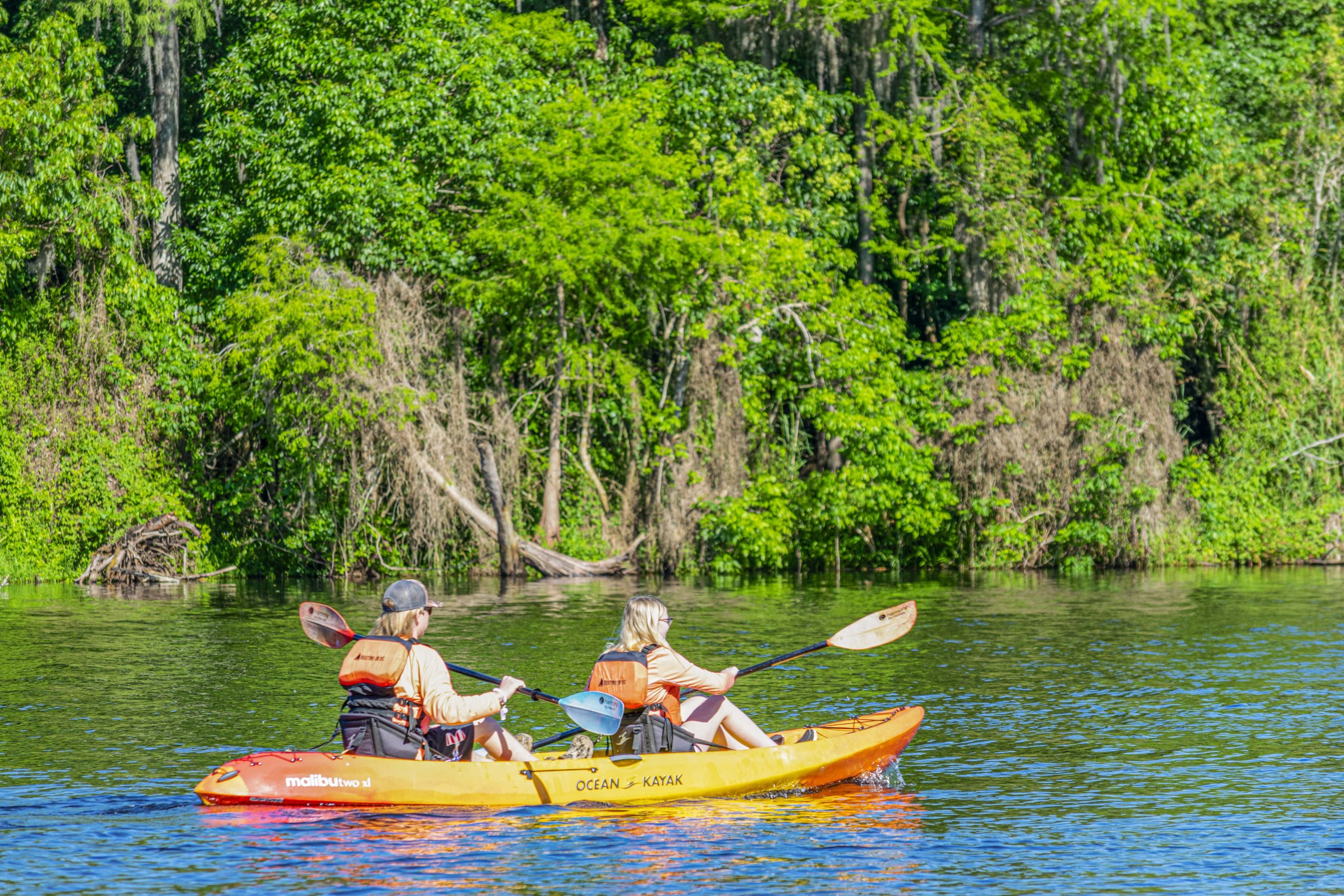 Two people paddling a yellow tandem kayak on a green-lined river.