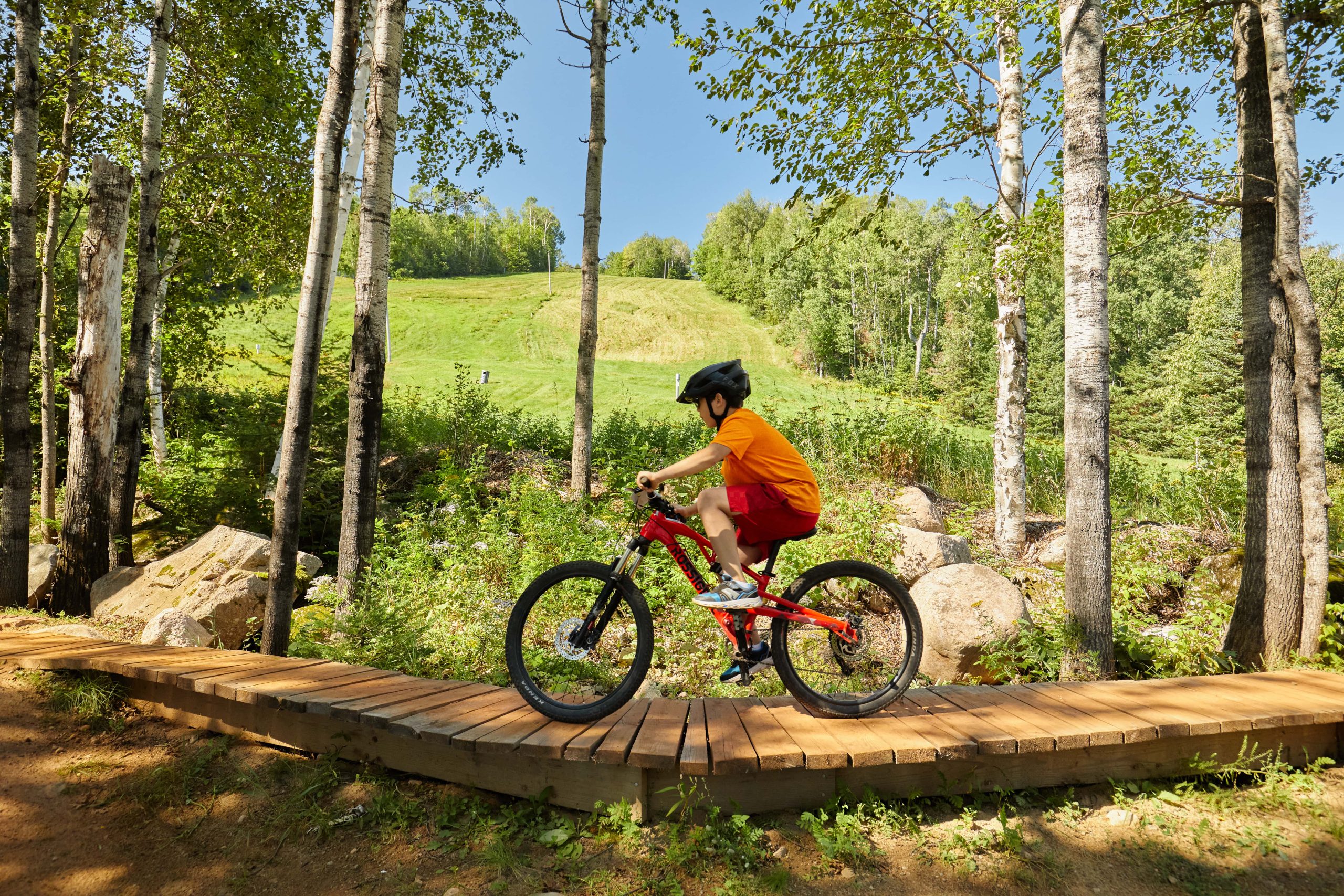 Boy biking on a wooden trail through forest at Giants Ridge Recreation Area, with a grassy hill and trees in the background.