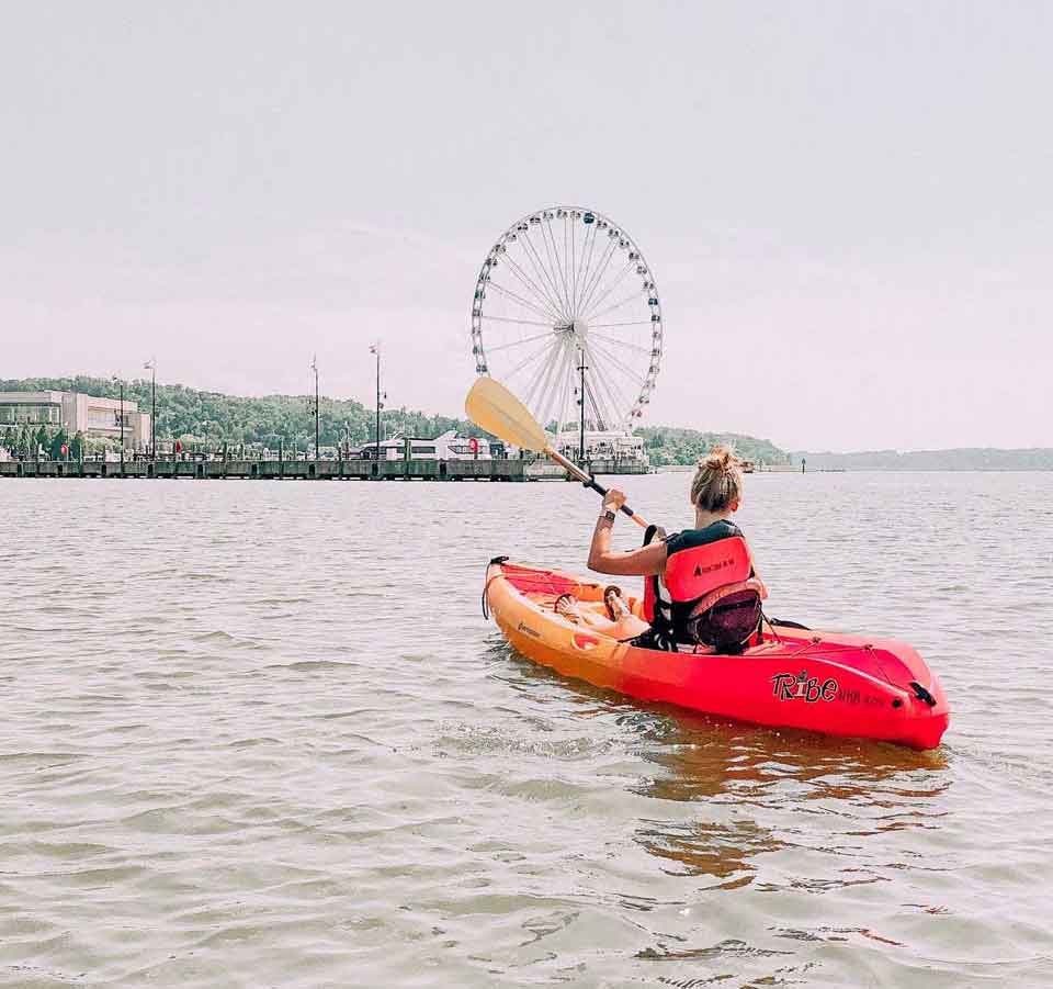 A woman paddles a red kayak toward a waterfront Ferris wheel, with calm water and distant buildings along the shore.